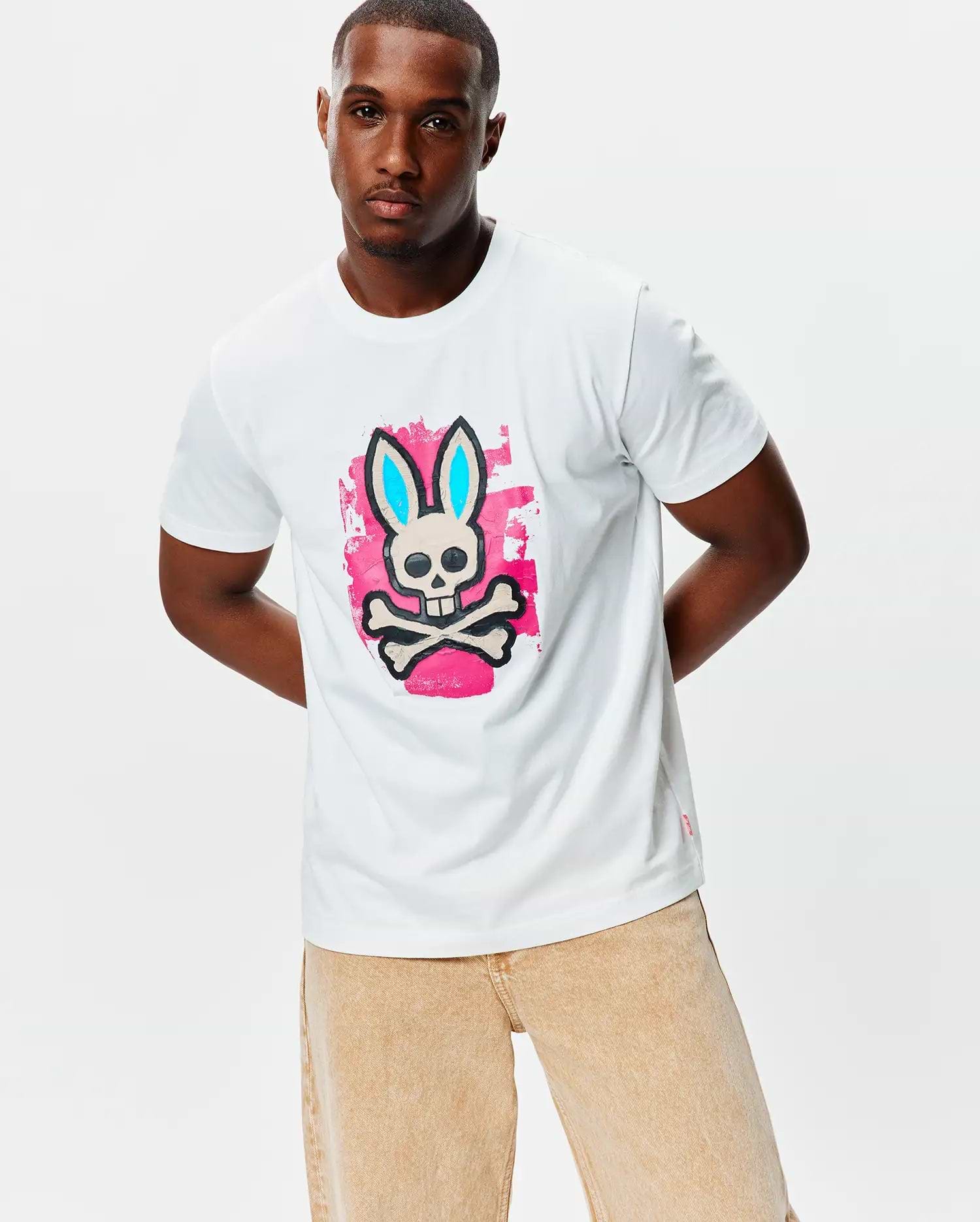 Mens Marcus Bunny Graphic Tee, a classic fit white jersey t-shirt featuring a large chest graphic of the Psycho Bunny skull with light blue ears on a pink paint splatter background, plus a small red hem tag.