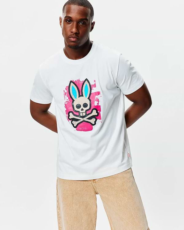 Mens Marcus Bunny Graphic Tee, a classic fit white jersey t-shirt featuring a large chest graphic of the Psycho Bunny skull with light blue ears on a pink paint splatter background, plus a small red hem tag.