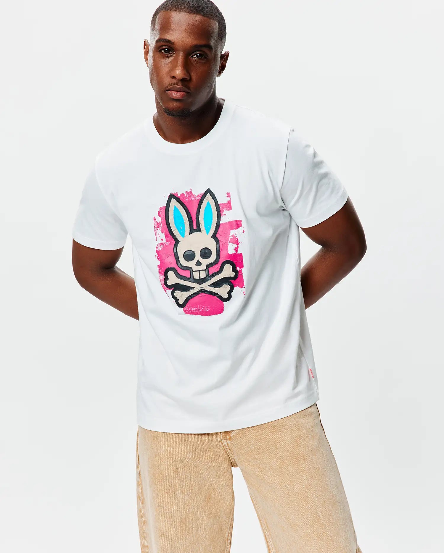 Mens Marcus Bunny Graphic Tee, a classic fit white jersey t-shirt featuring a large chest graphic of the Psycho Bunny skull with light blue ears on a pink paint splatter background, plus a small red hem tag.