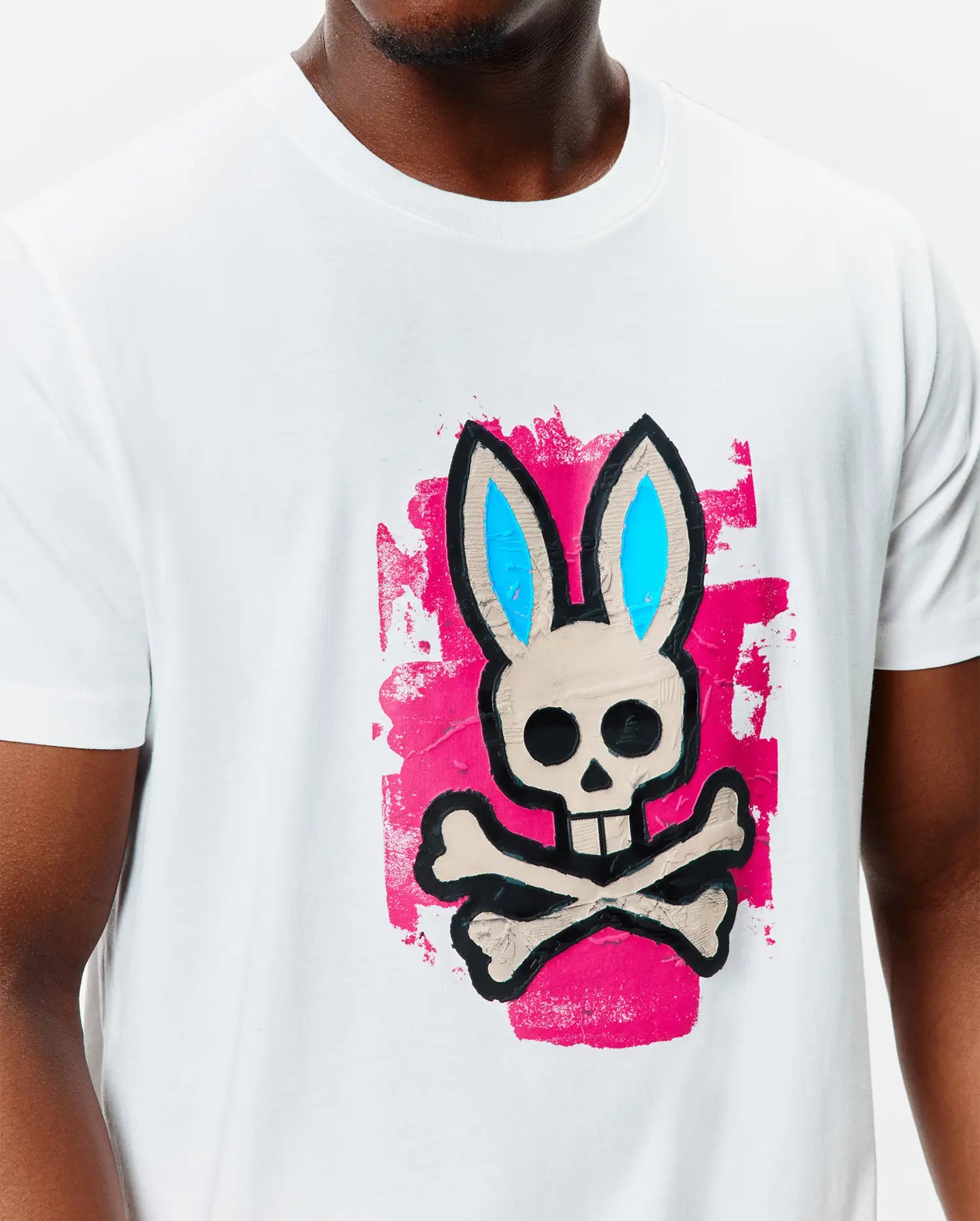 Mens Marcus Bunny Graphic Tee: White classic fit jersey t-shirt featuring a large central graphic of the Psycho Bunny skull and crossbones in beige, black, and bright blue, set against a distressed hot pink background.