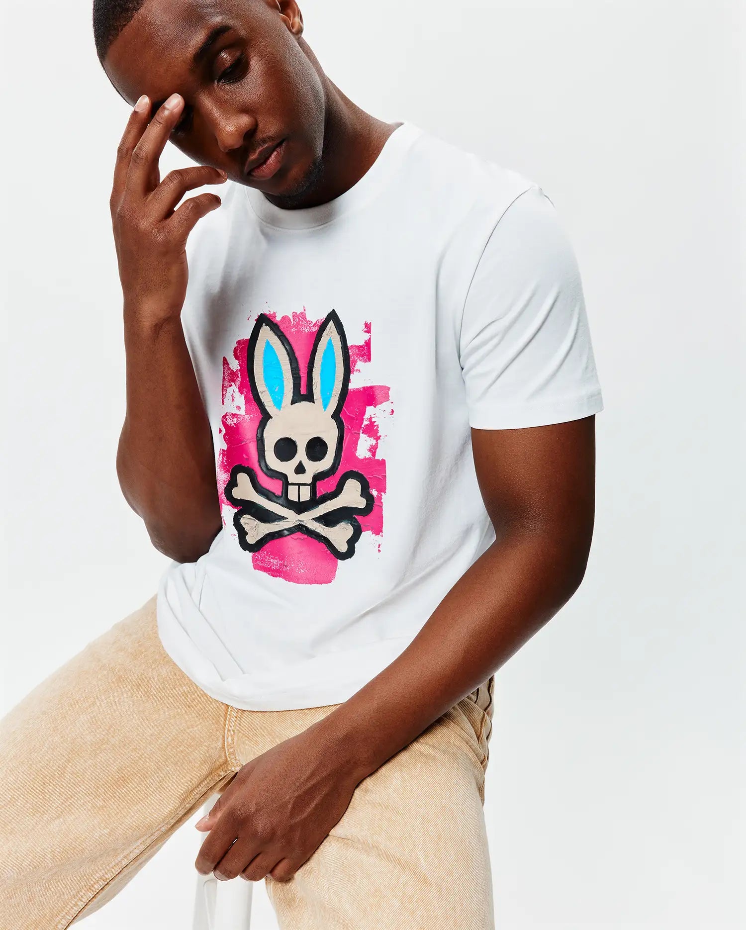 White Mens Marcus Bunny Graphic Tee, a classic fit jersey t-shirt featuring a large central Psycho Bunny skull and crossbones logo with blue ears over a vibrant pink brushstroke graphic.
