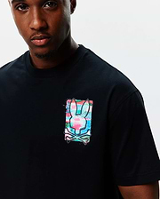 Mens Isaac Relaxed Fit Tee, black soft jersey with a relaxed fit, featuring a medium-sized left chest graphic of the Psycho Bunny skull logo in a vibrant pink, blue, and green paint splatter design.