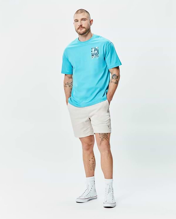 Mens Isaac Relaxed Fit Tee in aqua blue jersey, featuring a blue and white graphic print on the left chest, paired with light beige relaxed shorts with a small embroidered Psycho Bunny logo on the left thigh.