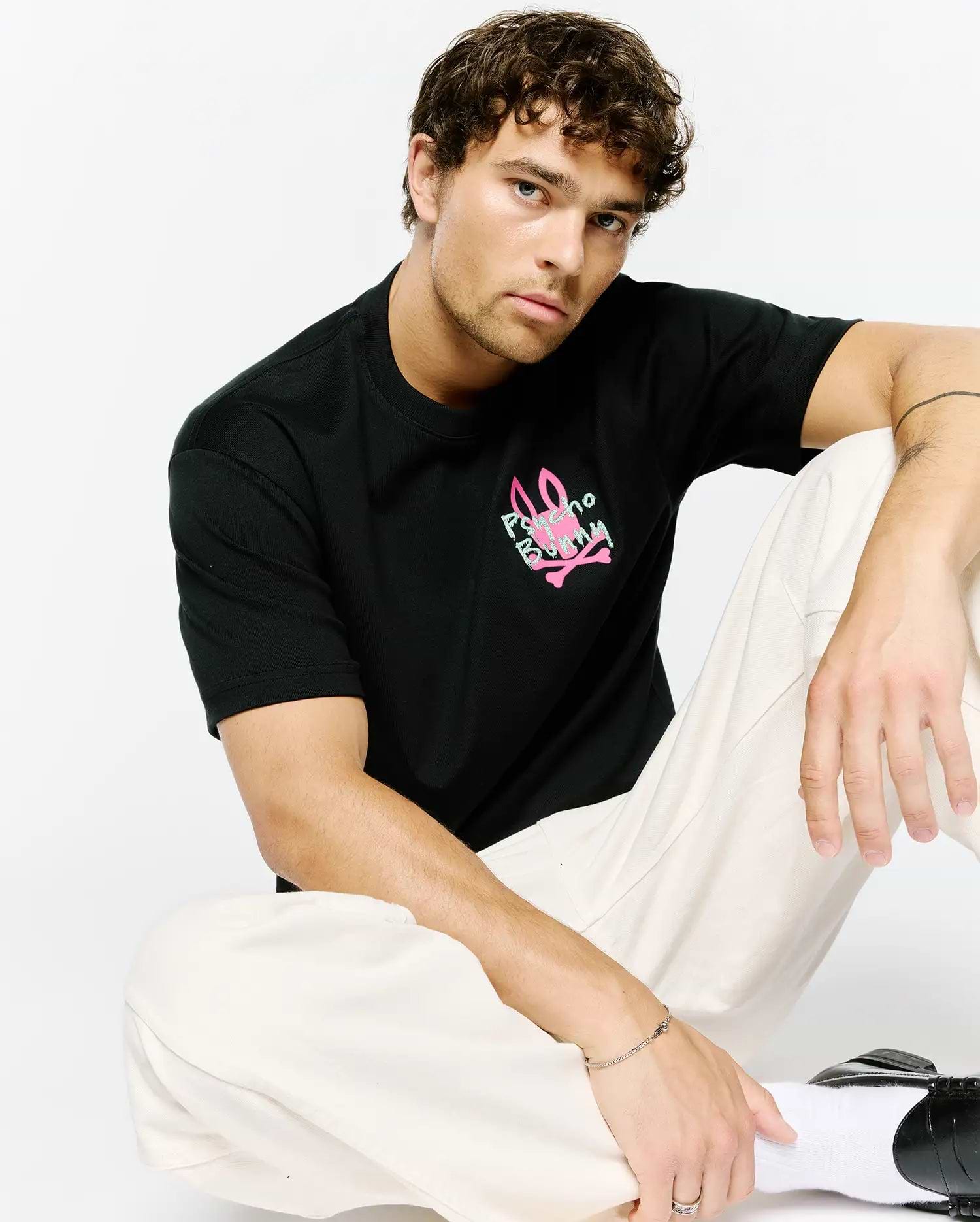 Mens Duncan Relaxed Fit Tee, a black jersey t-shirt with a relaxed fit, featuring a vibrant neon pink and light green Psycho Bunny skull logo graphic print on the chest.