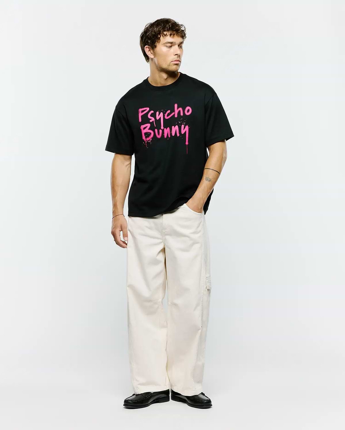 Mens Duncan Wordmark Relaxed Fit Tee, a black jersey t-shirt featuring a bold, large pink graffiti-style Psycho Bunny wordmark graphic with splatter details on the center chest.