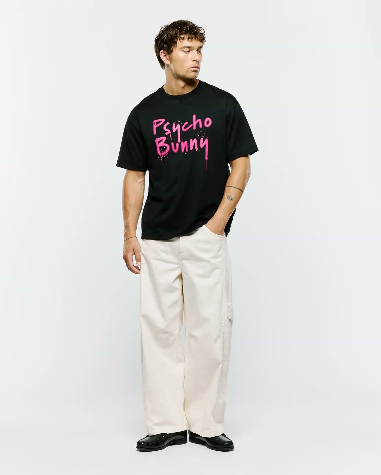 Mens Duncan Wordmark Relaxed Fit Tee, a black jersey t-shirt featuring a bold, large pink graffiti-style Psycho Bunny wordmark graphic with splatter details on the center chest.
