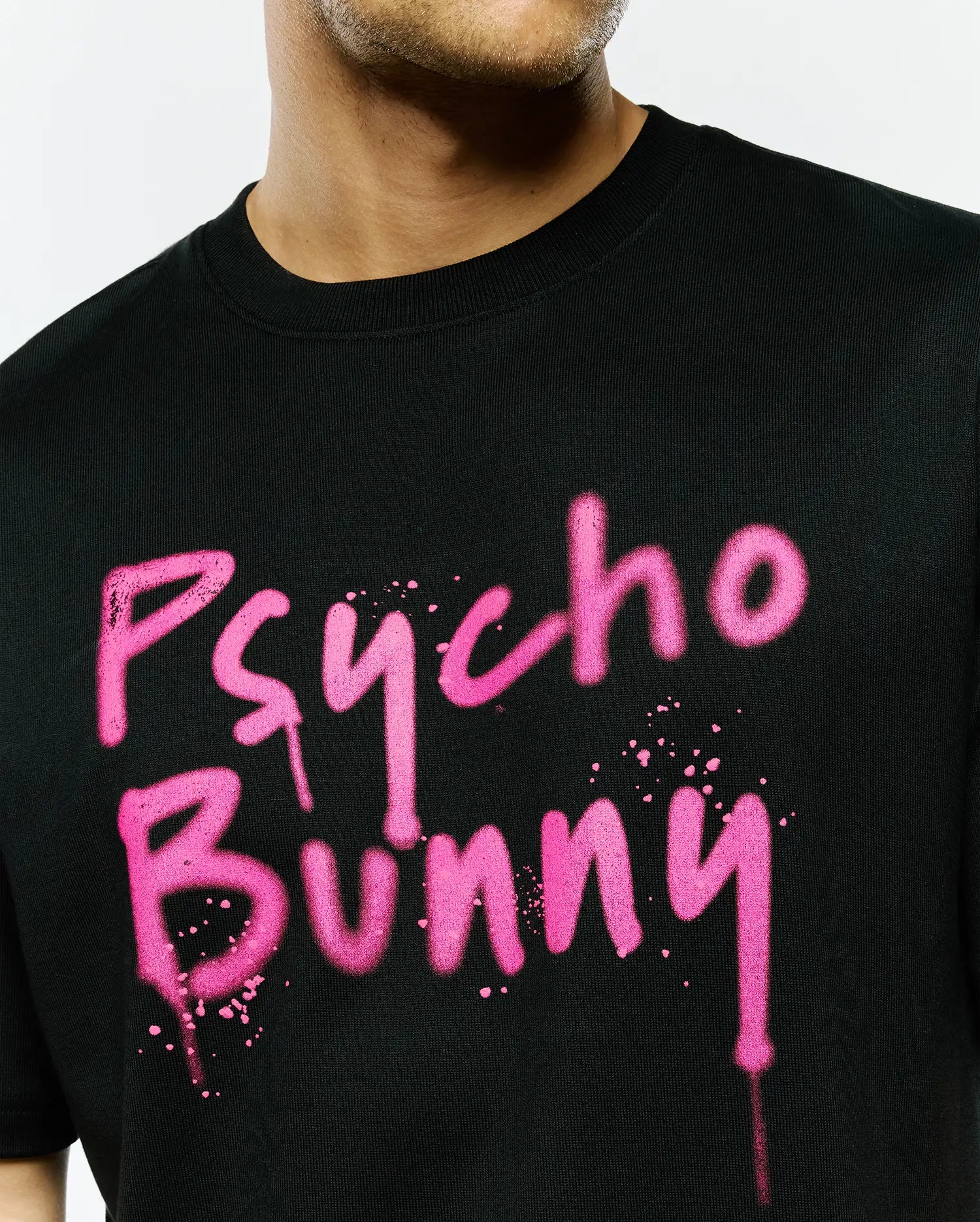Mens Duncan Wordmark Relaxed Fit Tee, black jersey knit with a large pink graffiti-style Psycho Bunny wordmark, featuring paint splatters and drips on the chest.