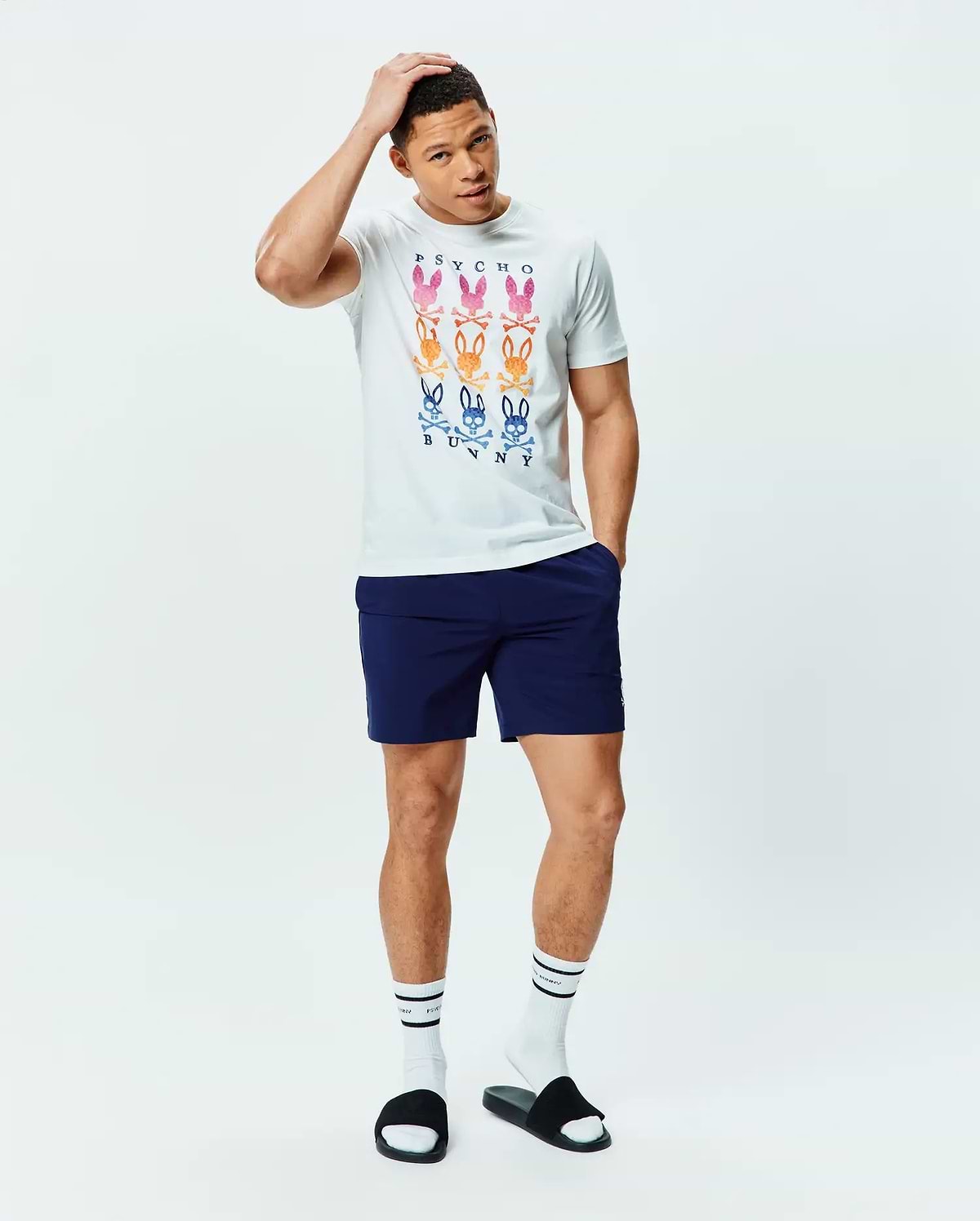 White Mens Colt Graphic Tee, classic fit, soft jersey, with a large central graphic of nine gradient multi-color Psycho Bunny skull logos and text, paired with navy athletic shorts featuring a small white Psycho Bunny logo.