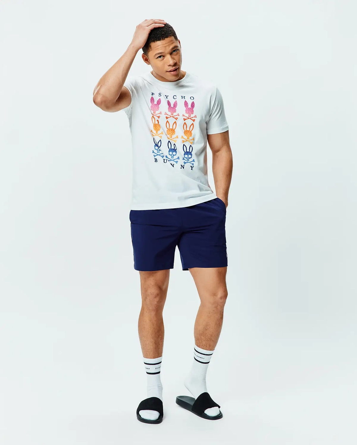 White Mens Colt Graphic Tee, classic fit, soft jersey, with a large central graphic of nine gradient multi-color Psycho Bunny skull logos and text, paired with navy athletic shorts featuring a small white Psycho Bunny logo.