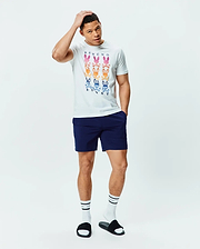 White Mens Colt Graphic Tee, classic fit, soft jersey, with a large central graphic of nine gradient multi-color Psycho Bunny skull logos and text, paired with navy athletic shorts featuring a small white Psycho Bunny logo.