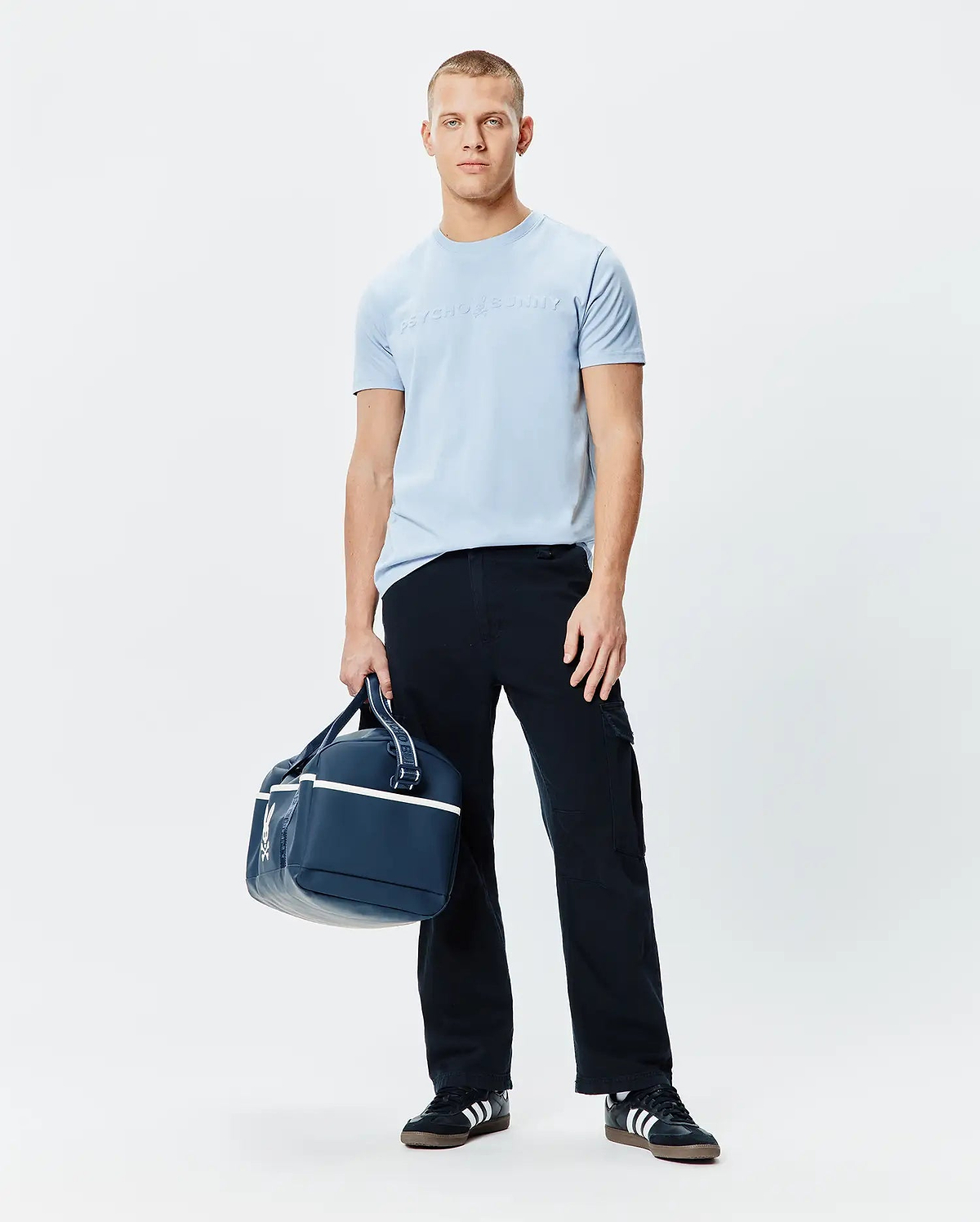 Mens Royce Graphic Tee, a light blue classic fit jersey t-shirt featuring a large, subtle tone-on-tone embossed Psycho Bunny text logo prominently centered on the chest.