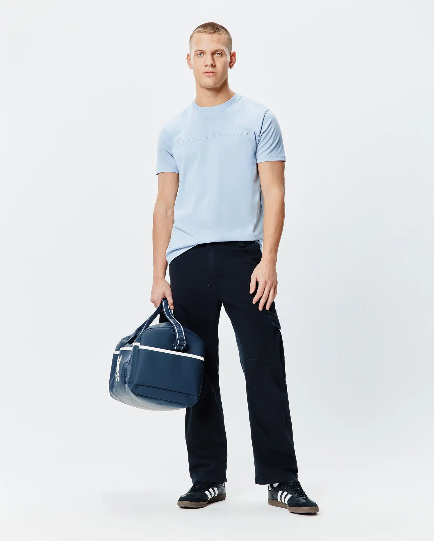 Mens Royce Graphic Tee, a light blue classic fit jersey t-shirt featuring a large, subtle tone-on-tone embossed Psycho Bunny text logo prominently centered on the chest.