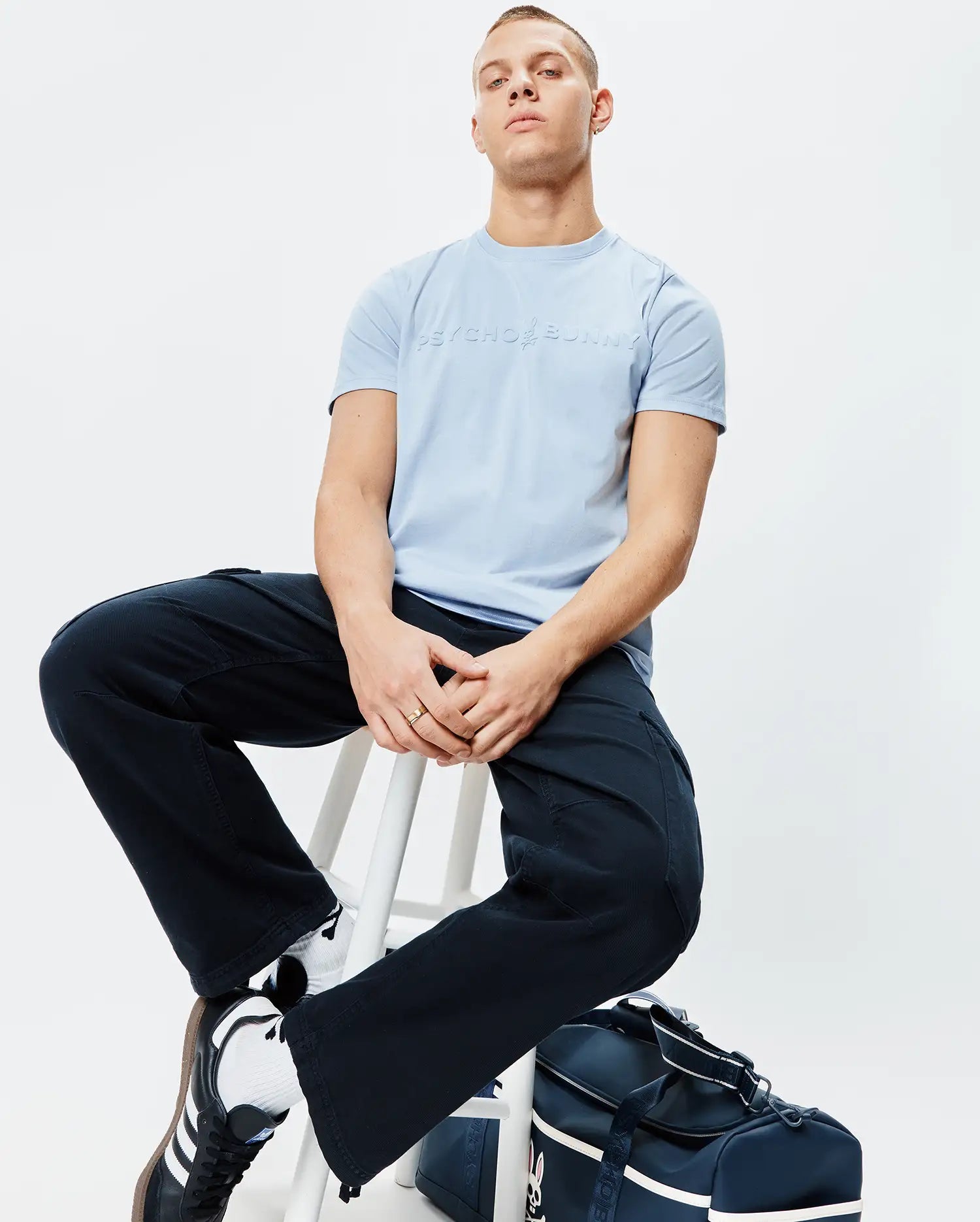 Light blue Mens Royce Graphic Tee, a classic fit, smooth jersey knit, featuring a subtle tone-on-tone embossed Psycho Bunny logo and text centered on the chest.