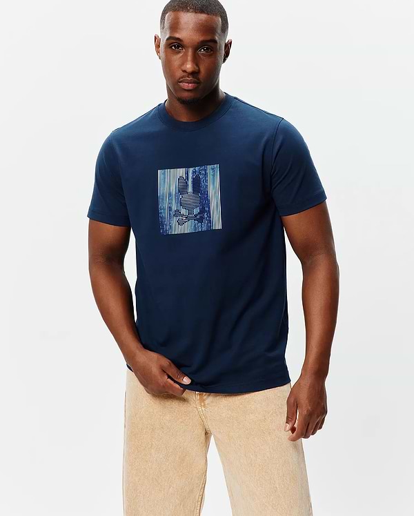Mens Seamus Graphic Tee, a navy blue classic fit jersey t-shirt featuring a large chest graphic of the Psycho Bunny skull logo within a vibrant blue and white striped rectangle.