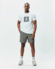 Mens Langston Graphic Tee, an off-white jersey tee with a large, central glitch-effect Psycho Bunny skull graphic in pink, blue, and black stripes, paired with olive green relaxed fit shorts.