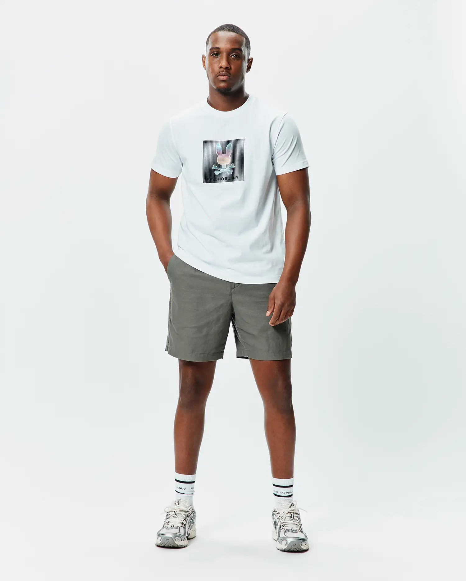 Mens Langston Graphic Tee, an off-white jersey tee with a large, central glitch-effect Psycho Bunny skull graphic in pink, blue, and black stripes, paired with olive green relaxed fit shorts.