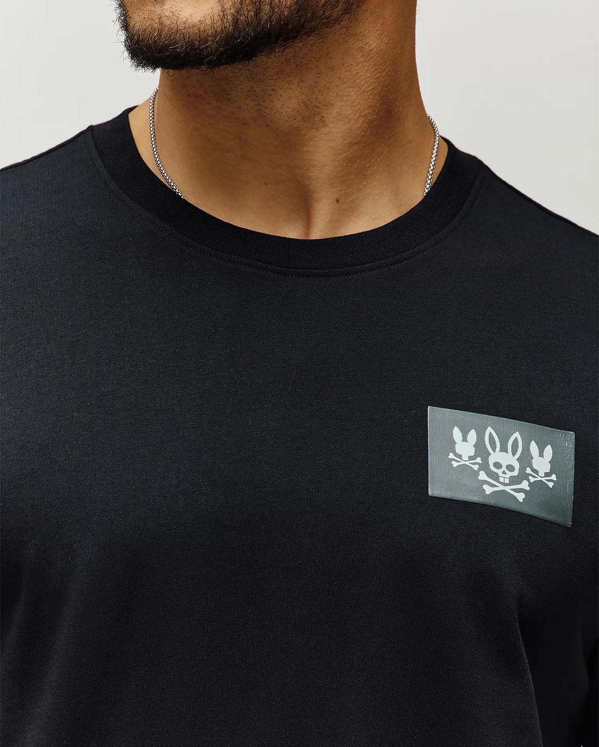 Mens Jarvis Chest Graphic Tee, a black classic fit premium jersey knit with a bold rectangular chest patch featuring three white Psycho Bunny skull and crossbones logos.