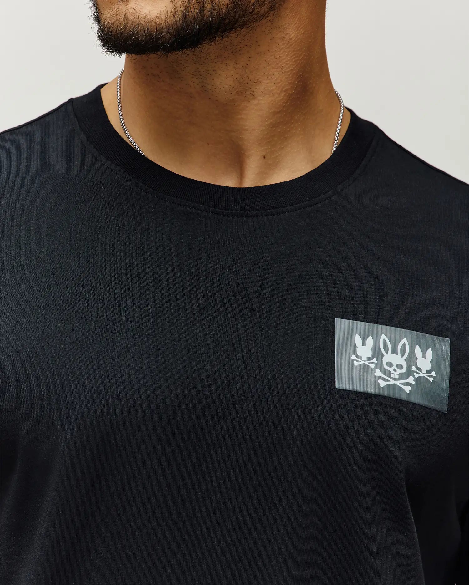 Mens Jarvis Chest Graphic Tee, a black classic fit premium jersey knit with a bold rectangular chest patch featuring three white Psycho Bunny skull and crossbones logos.
