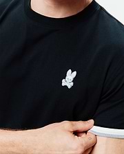 Mens Holden Tee, black classic fit jersey knit with a small embroidered white Psycho Bunny skull logo on the left chest, featuring a ribbed collar and striped white and grey sleeve cuffs.