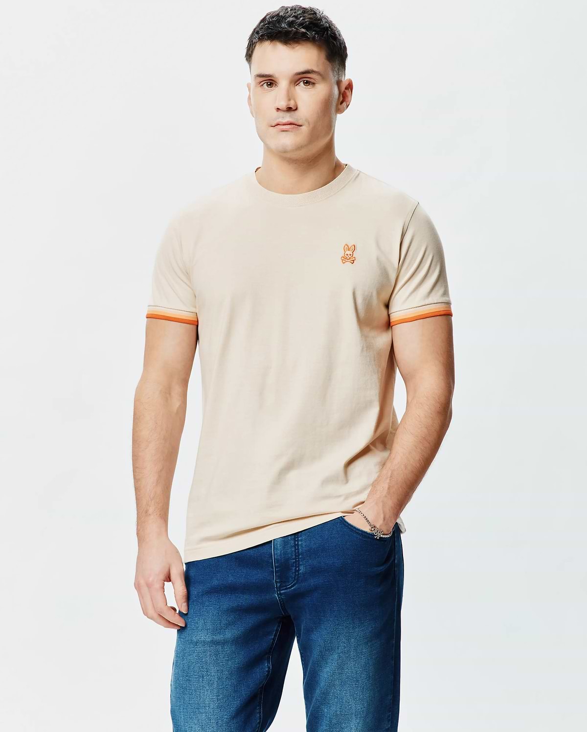 Mens Holden Tee, a classic fit beige jersey t-shirt featuring a small orange embroidered Psycho Bunny skull logo on the left chest and orange striped sleeve cuffs.