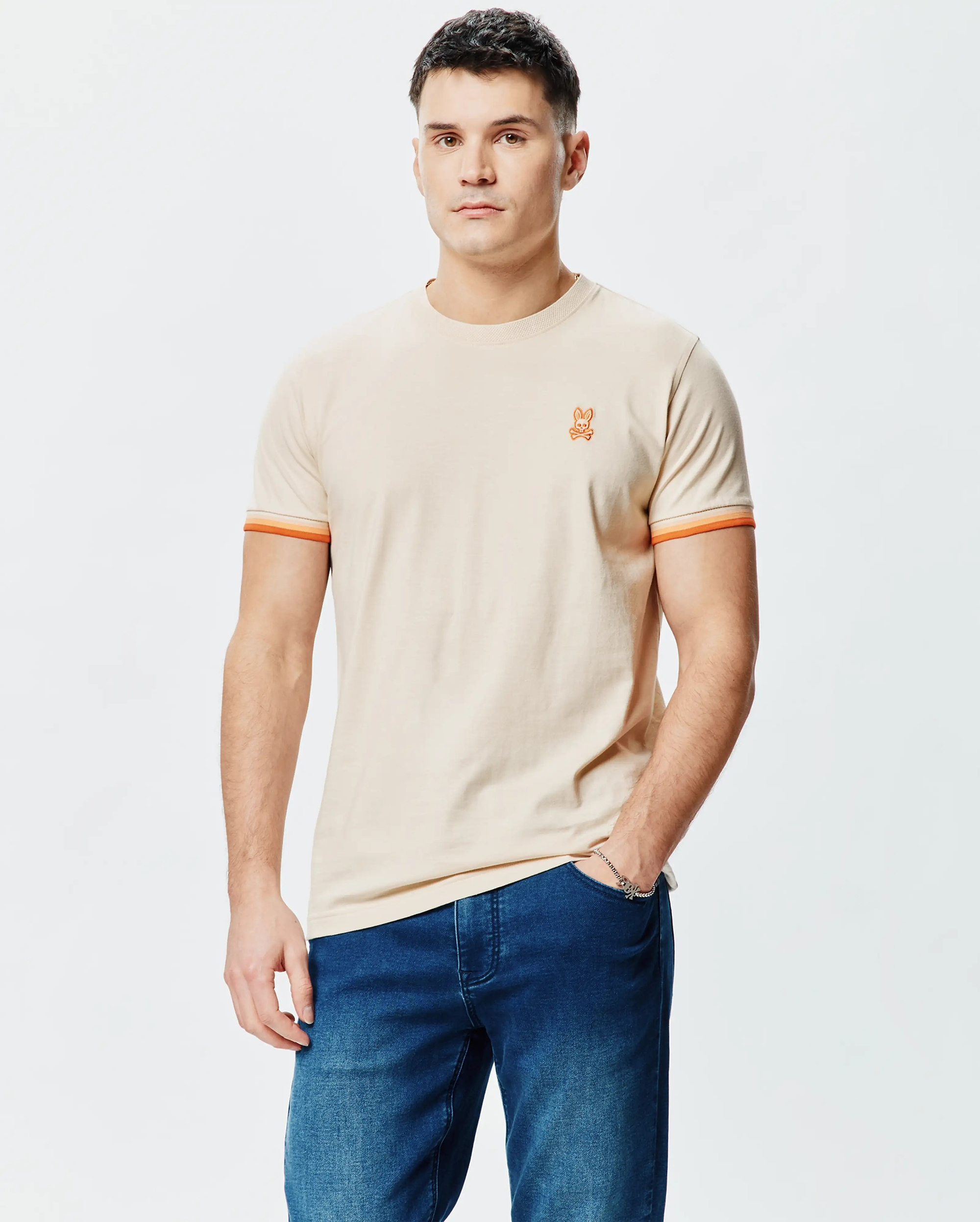 Mens Holden Tee, a classic fit beige jersey t-shirt featuring a small orange embroidered Psycho Bunny skull logo on the left chest and orange striped sleeve cuffs.