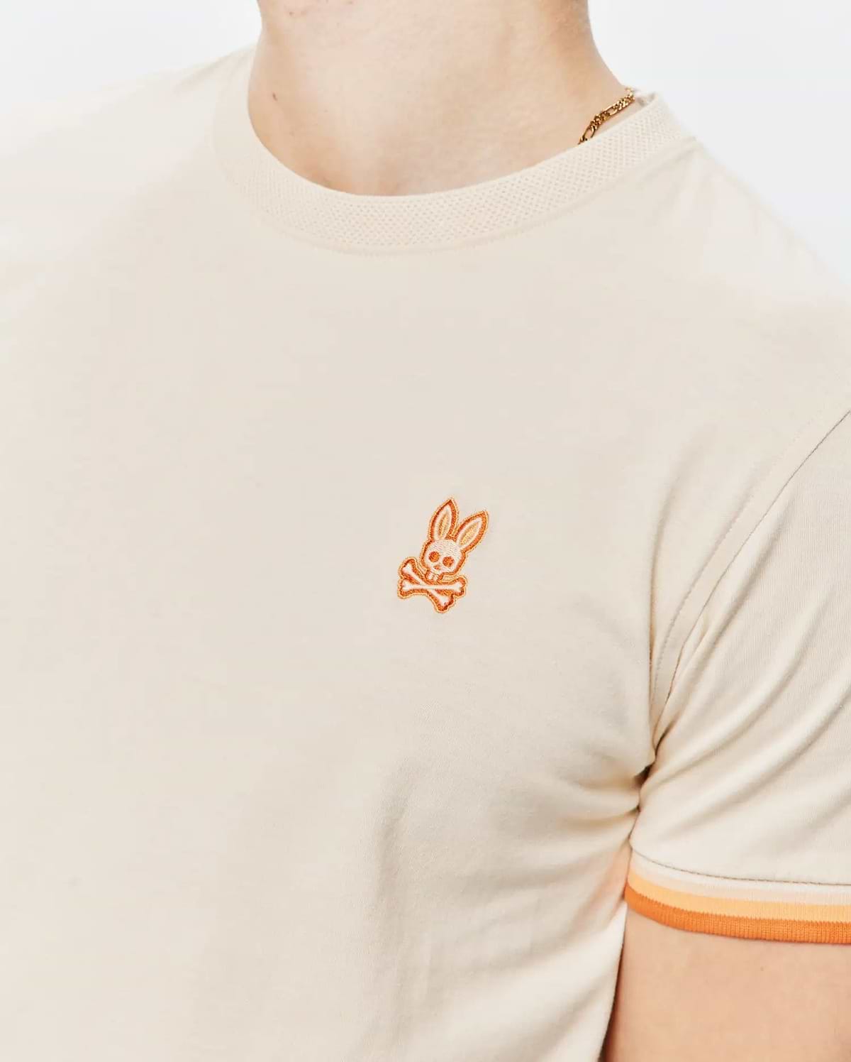 Mens Holden Tee in cream jersey knit with a ribbed collar, featuring a small embroidered orange Psycho Bunny skull logo on the left chest and ribbed sleeve cuffs with orange and peach stripes.