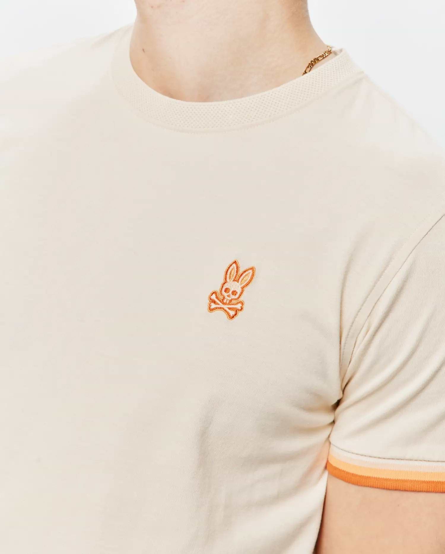 Mens Holden Tee in cream jersey knit with a ribbed collar, featuring a small embroidered orange Psycho Bunny skull logo on the left chest and ribbed sleeve cuffs with orange and peach stripes.