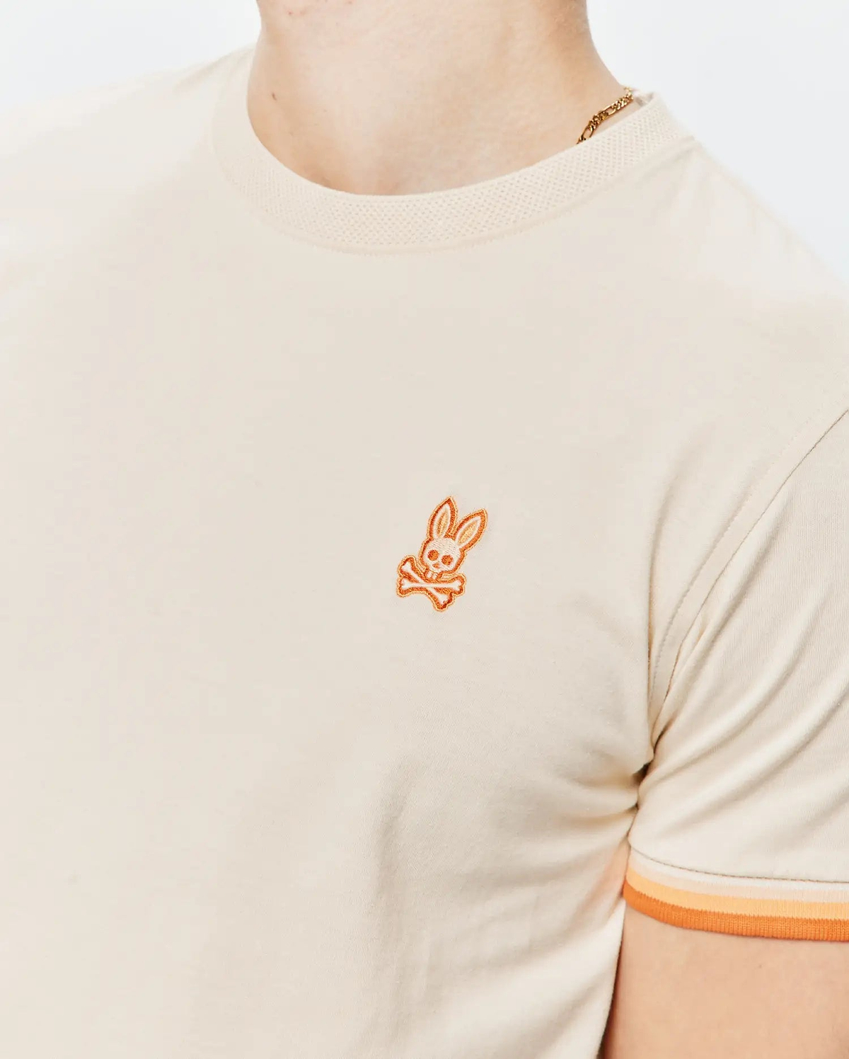 Mens Holden Tee in cream jersey knit with a ribbed collar, featuring a small embroidered orange Psycho Bunny skull logo on the left chest and ribbed sleeve cuffs with orange and peach stripes.