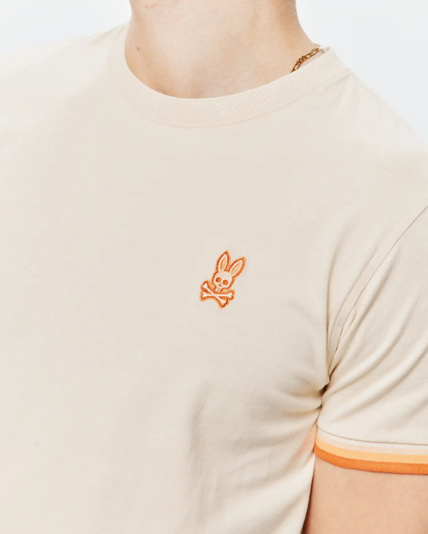 Mens Holden Tee in cream jersey knit with a ribbed collar, featuring a small embroidered orange Psycho Bunny skull logo on the left chest and ribbed sleeve cuffs with orange and peach stripes.