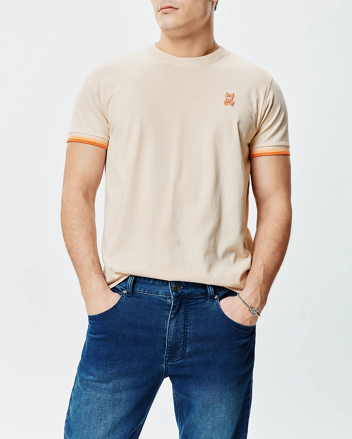 Mens Holden Tee, classic fit, beige jersey with small embroidered orange Psycho Bunny skull logo on left chest and orange striped sleeve cuffs.