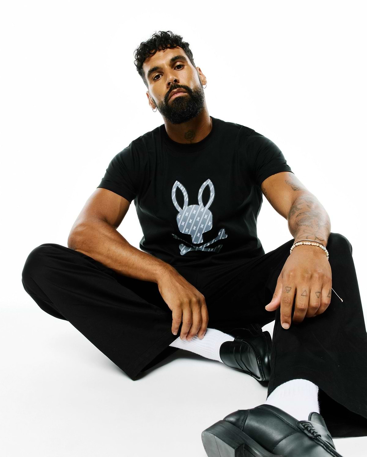 Mens Jaden Appliqué Graphic Tee: Black classic fit jersey t-shirt featuring a large central grey and white Psycho Bunny skull logo with a repeating mini-logo pattern.