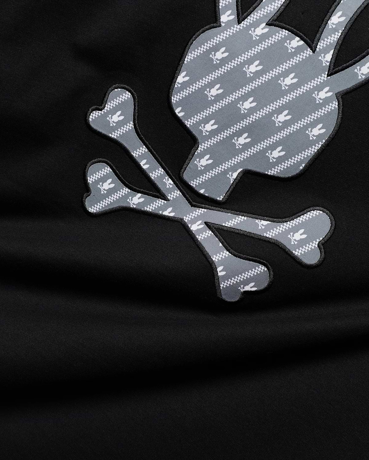 Mens Jaden Appliqué Graphic Tee. Black jersey with large grey and white Psycho Bunny skull and crossbones appliqué, featuring a repeating pattern of small white bunny skulls and chevron stripes.
