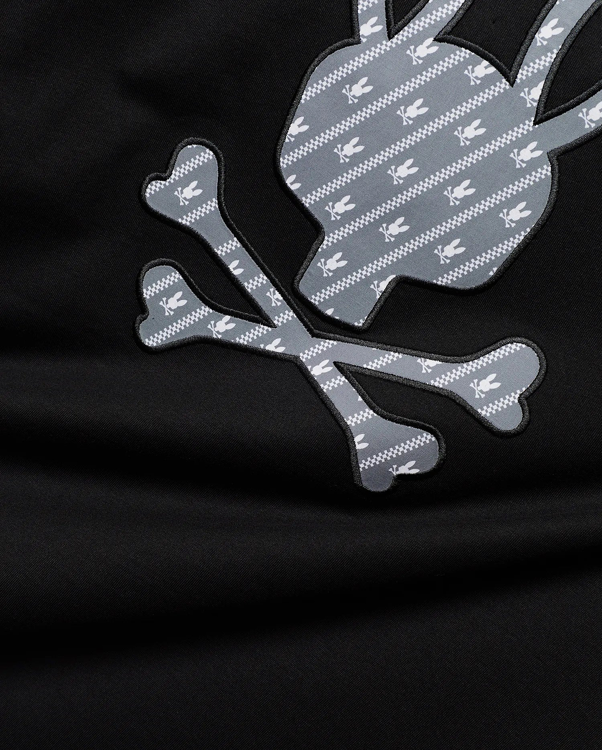 Mens Jaden Appliqué Graphic Tee. Black jersey with large grey and white Psycho Bunny skull and crossbones appliqué, featuring a repeating pattern of small white bunny skulls and chevron stripes.