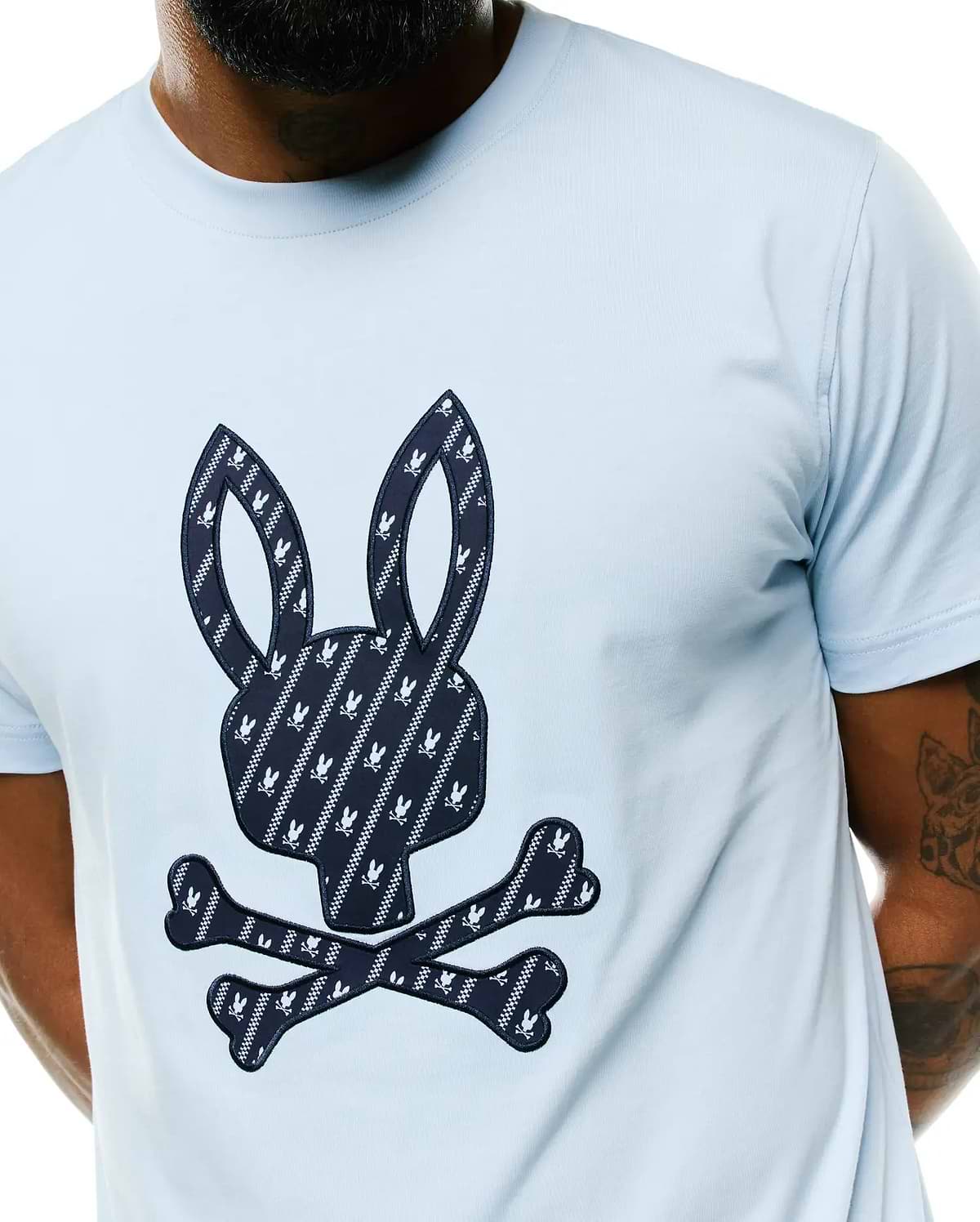 Light blue Mens Jaden Appliqué Graphic Tee, classic fit, smooth jersey fabric. Features a large navy and white patterned Psycho Bunny skull logo appliqué on the chest with small bunny skulls and crossbones.