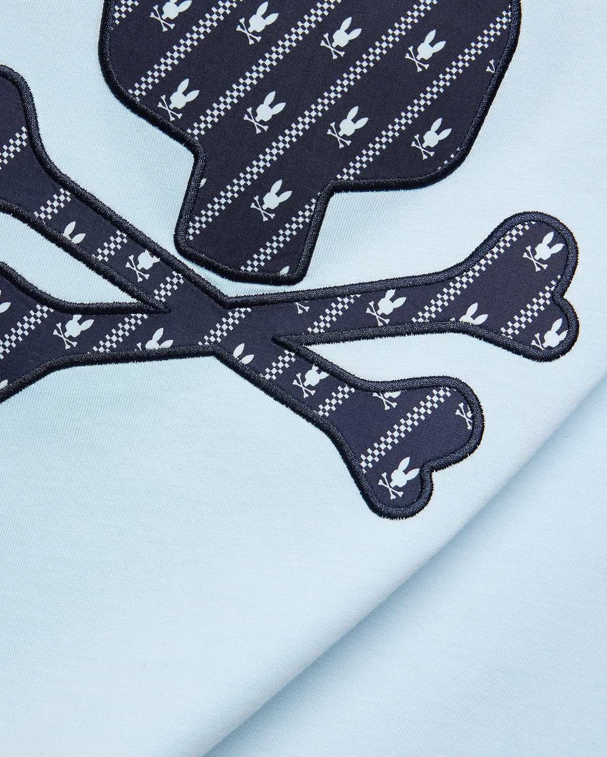 Mens Jaden Graphic Tee in light blue jersey knit, featuring a large navy blue skull and crossbones graphic with white Psycho Bunny logo and checkered stripe pattern, outlined in black embroidery.