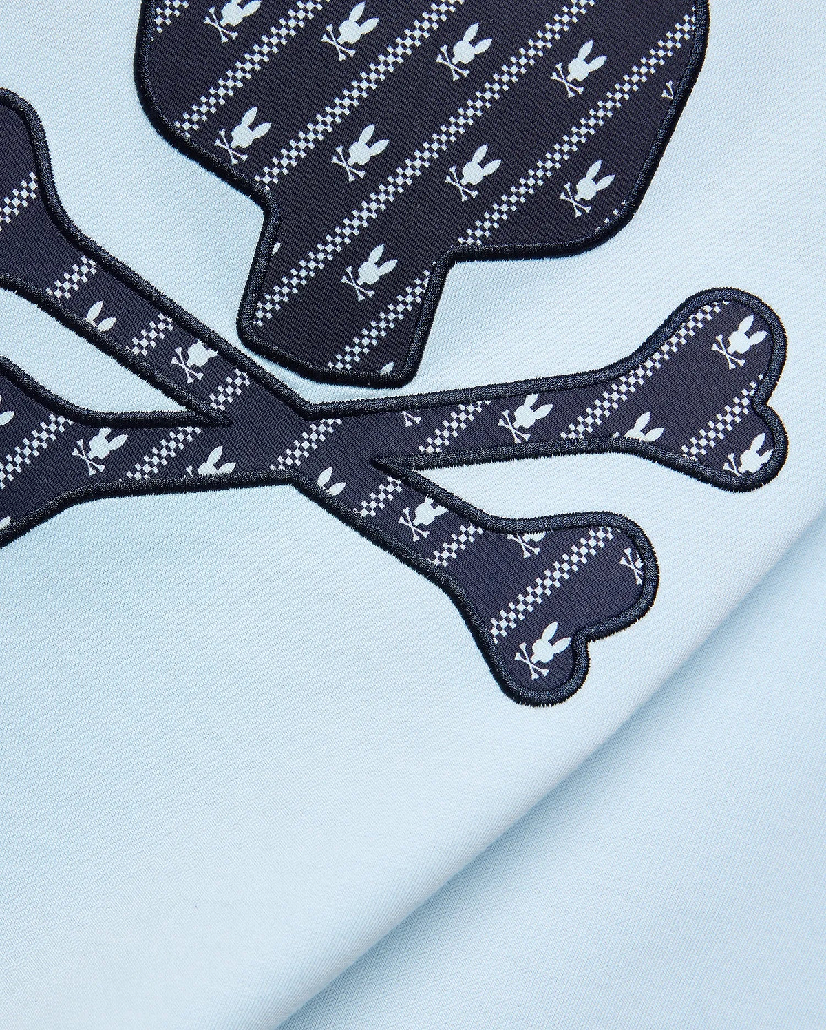 Mens Jaden Graphic Tee in light blue jersey knit, featuring a large navy blue skull and crossbones graphic with white Psycho Bunny logo and checkered stripe pattern, outlined in black embroidery.