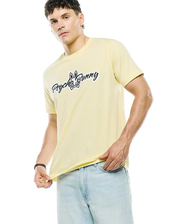Mens Jaydon Wordmark Graphic Tee, a classic fit pale yellow jersey t-shirt featuring a large black and white Psycho Bunny wordmark and skull logo graphic on the chest.
