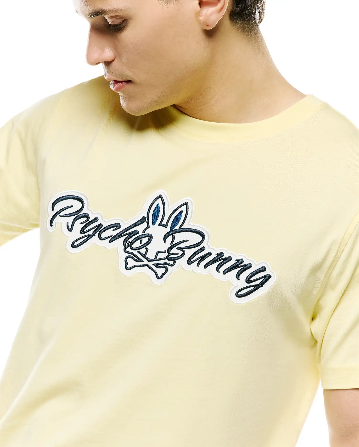 Mens Jaydon Wordmark Graphic Tee, a pale yellow jersey knit t-shirt featuring a large central embroidered Psycho Bunny wordmark and skull logo in navy and white.