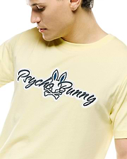 Mens Jaydon Wordmark Graphic Tee, a pale yellow jersey knit t-shirt featuring a large central embroidered Psycho Bunny wordmark and skull logo in navy and white.