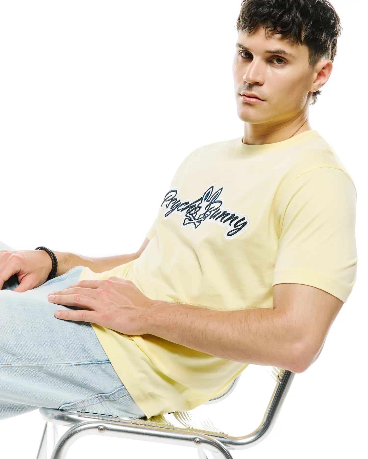 Mens Jaydon Wordmark Graphic Tee, a pale yellow classic fit jersey knit t-shirt featuring a prominent dark blue Psycho Bunny wordmark with integrated bunny skull logo, outlined in white, centered on the chest.