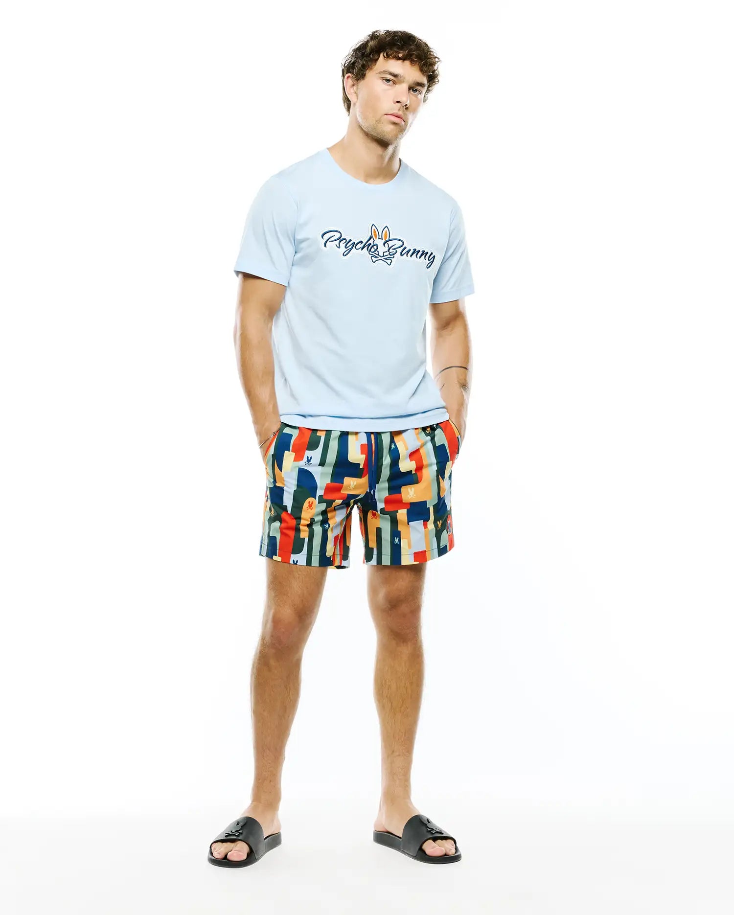 Mens Jaydon Wordmark Graphic Tee in light blue jersey, featuring a large blue Psycho Bunny wordmark and bunny skull logo on the chest, paired with multi-color abstract pattern shorts with subtle bunny skull logos.