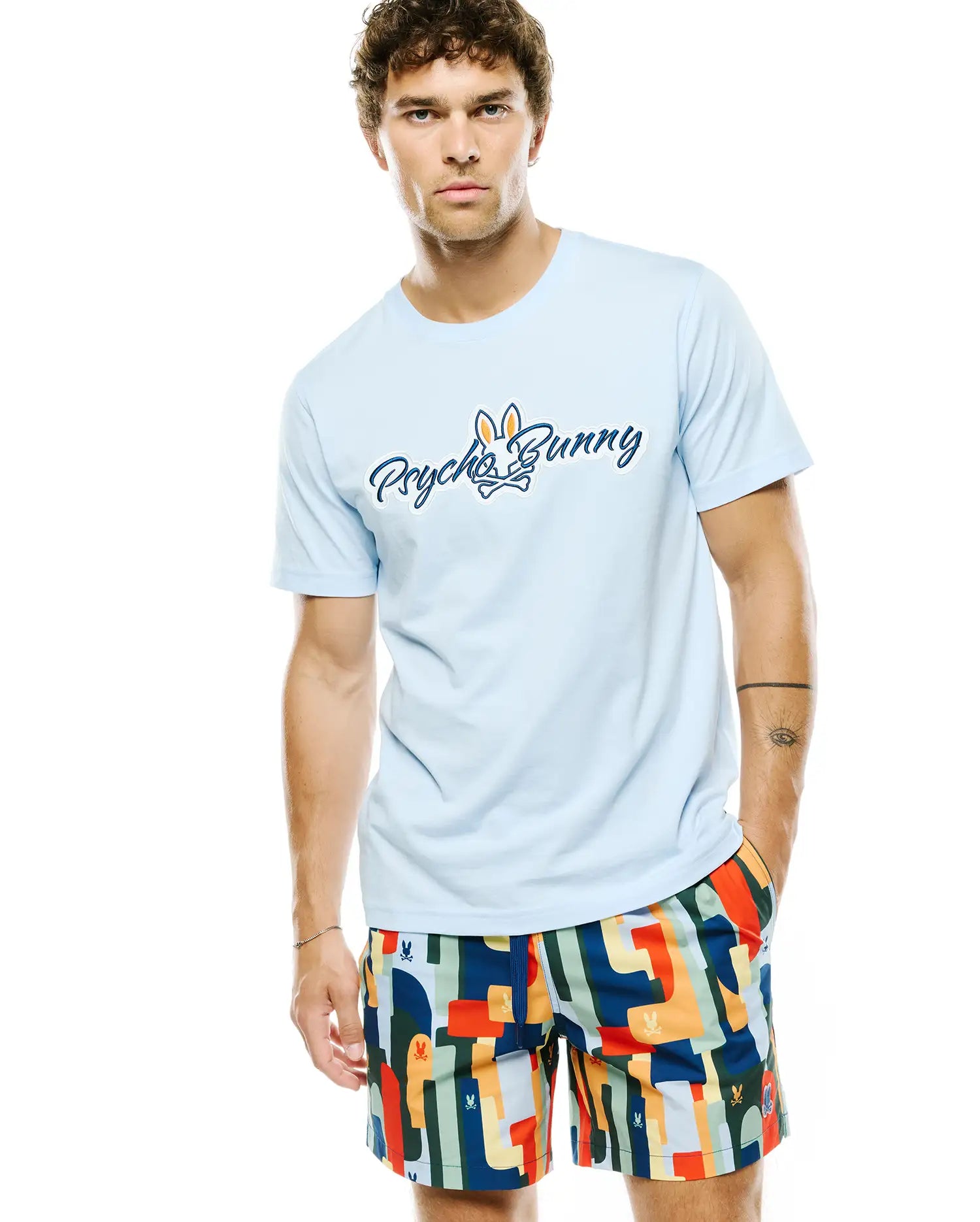 Mens Jaydon Wordmark Graphic Tee, a light blue classic fit jersey t-shirt featuring a large blue script Psycho Bunny wordmark and orange and white bunny skull logo on the chest.