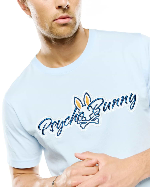 Mens Jaydon Wordmark Graphic Tee, light blue classic fit jersey knit t-shirt with a large embroidered chest graphic: blue Psycho Bunny wordmark and skull logo with white outline and orange ear details.