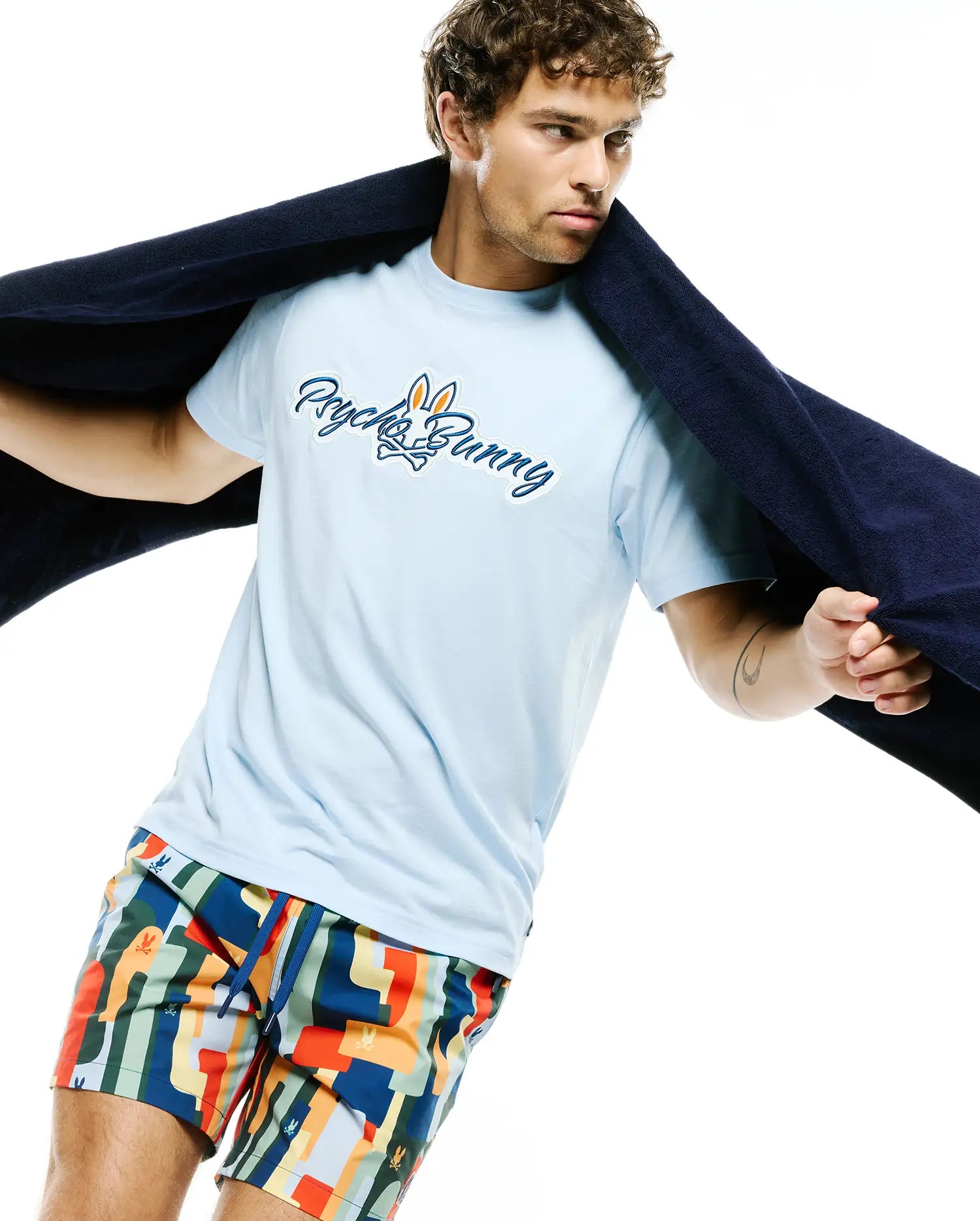 Light blue Mens Jaydon Wordmark Graphic Tee with a large embroidered Psycho Bunny wordmark and skull logo on the chest. Multi-color geometric pattern swim shorts feature small repeated bunny skull logos.