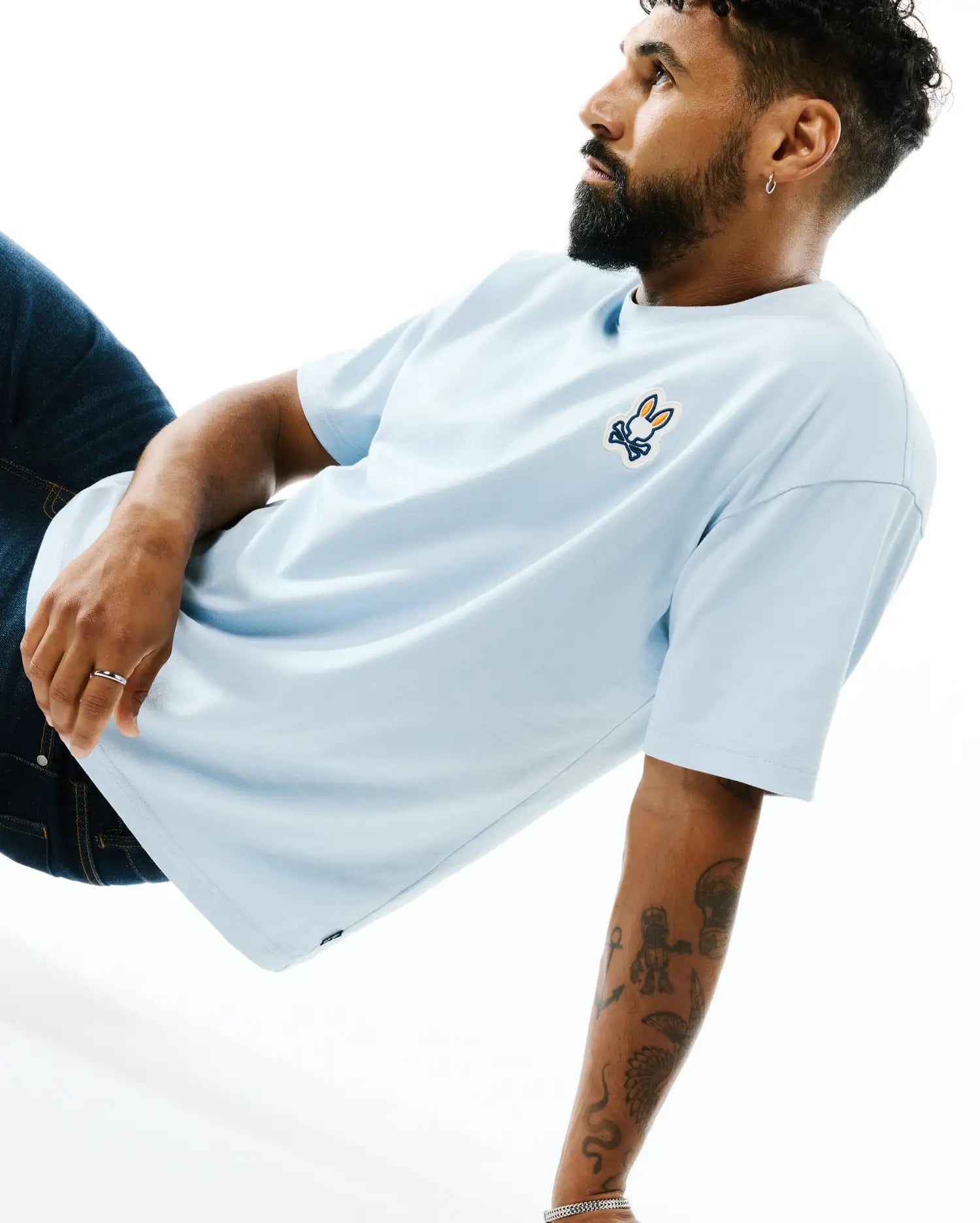 Mens Jaydon Pima Cotton Oversized-Fit Tee in light blue, featuring a smooth jersey texture and a vibrant orange, white, and blue embroidered Psycho Bunny skull logo on the left chest.