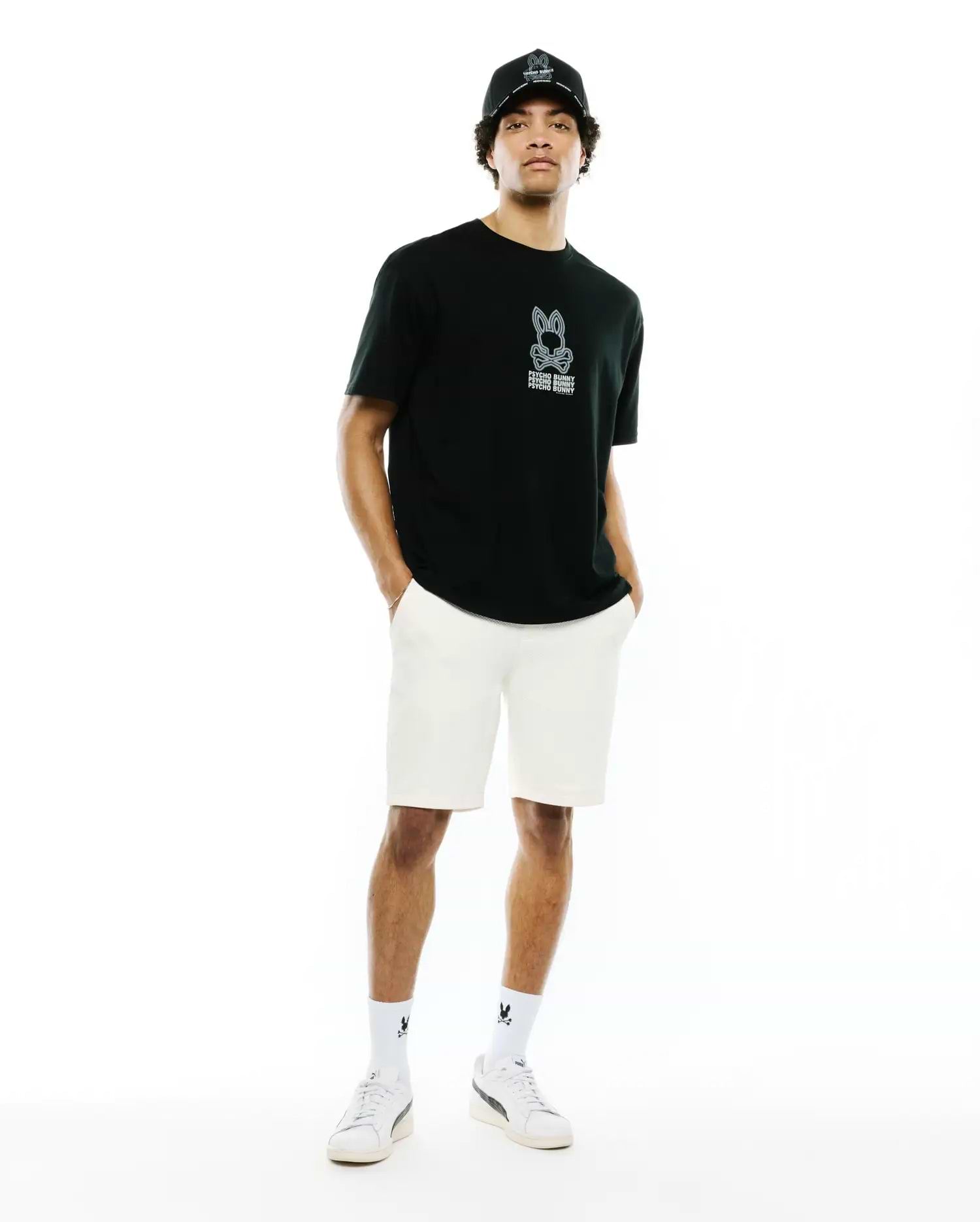 Mens Alex Relaxed Back Graphic Tee in black jersey, featuring a light blue outlined Psycho Bunny skull logo and white text on the left chest, paired with cream shorts, a black logo cap, and white sneakers.