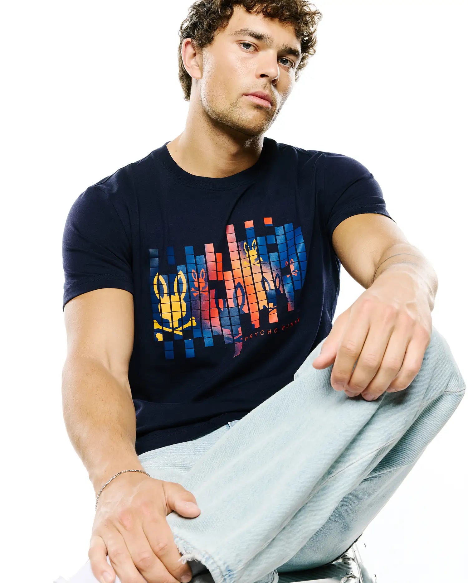 Mens Zander Graphic Tee, a classic fit navy blue jersey t-shirt featuring a large pixelated chest graphic with the iconic Psycho Bunny skull logo and brand name in vibrant orange, yellow, and blue hues.