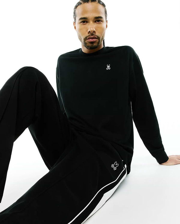 Black Mens Norman Long Sleeve Oversized Fit Tee with a white Psycho Bunny skull logo on the left chest, and black track pants with a white side stripe and a thigh bunny logo.