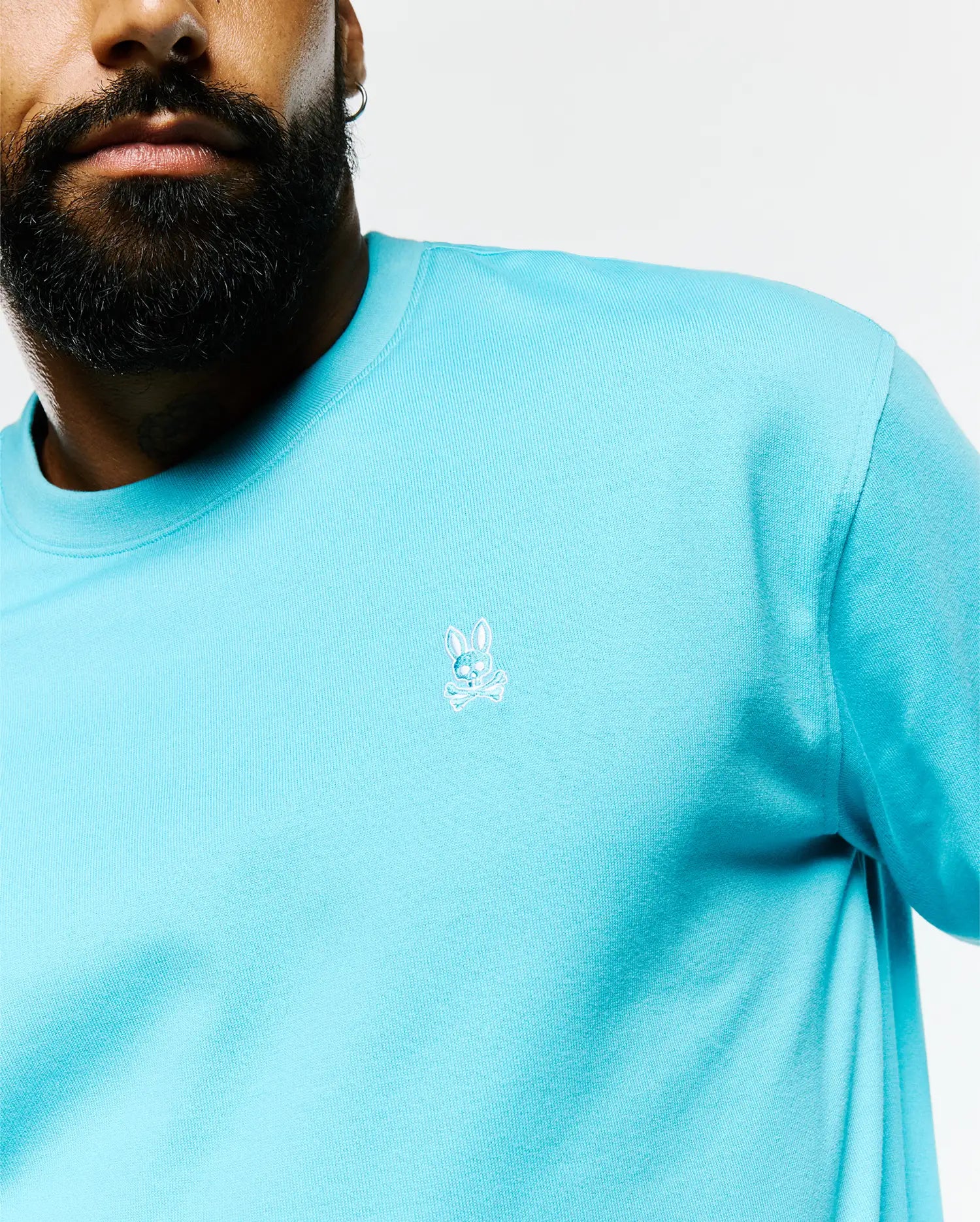 Mens Norman Long Sleeve Relaxed Fit Tee in vibrant aqua jersey knit, featuring a small, intricately embroidered white and light blue Psycho Bunny skull logo on the left chest.