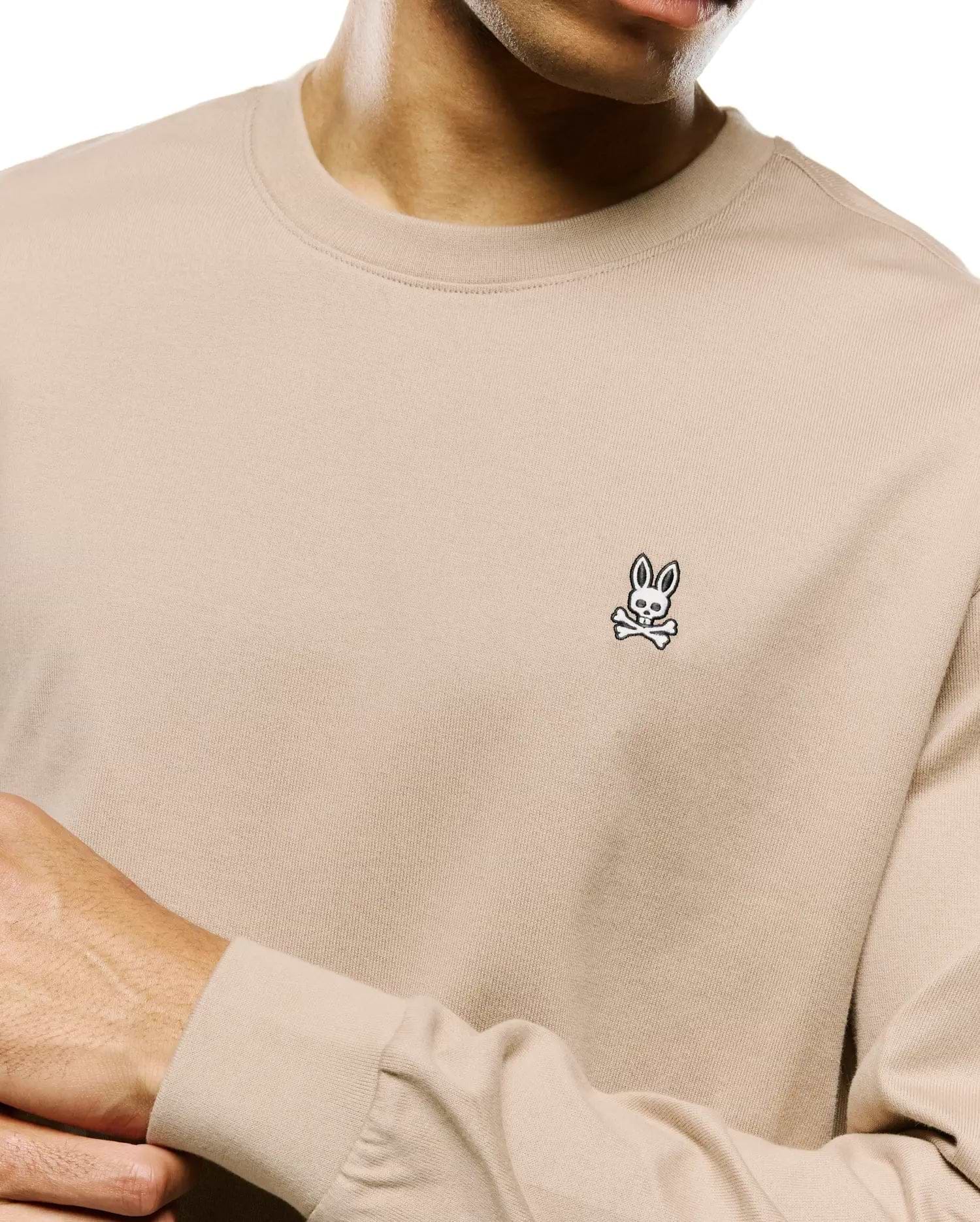 Mens Norman Long Sleeve Relaxed Fit Tee in beige, featuring a soft jersey knit texture and a small, embroidered black and white Psycho Bunny skull logo on the left chest.